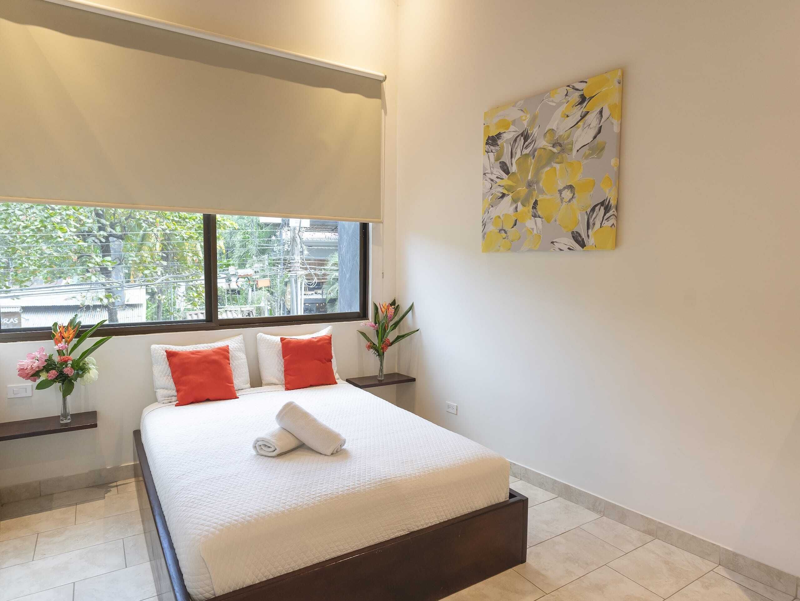 Un dormitorio moderno con un ventanal grande, una cama con ropa de cama blanca y almohadas naranjas, una mesita de noche con un florero y un cuadro en la pared.