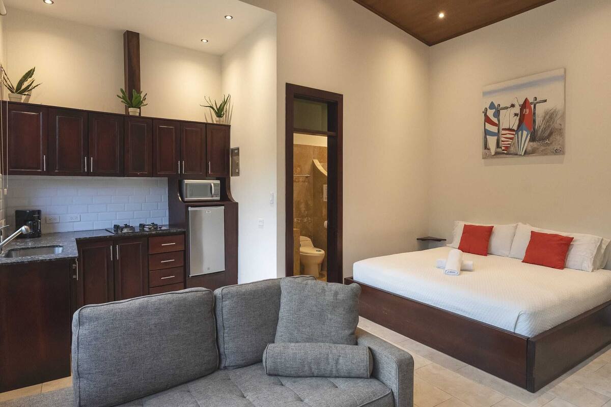 A modern bedroom with a large bed, a grey sofa, and a kitchen area with dark wooden cabinets and a microwave.