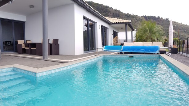 Une piscine extérieure moderne, avec une eau d’un bleu limpide, entourée d’un bâtiment blanc doté de grandes fenêtres et d’un espace détente abrité.