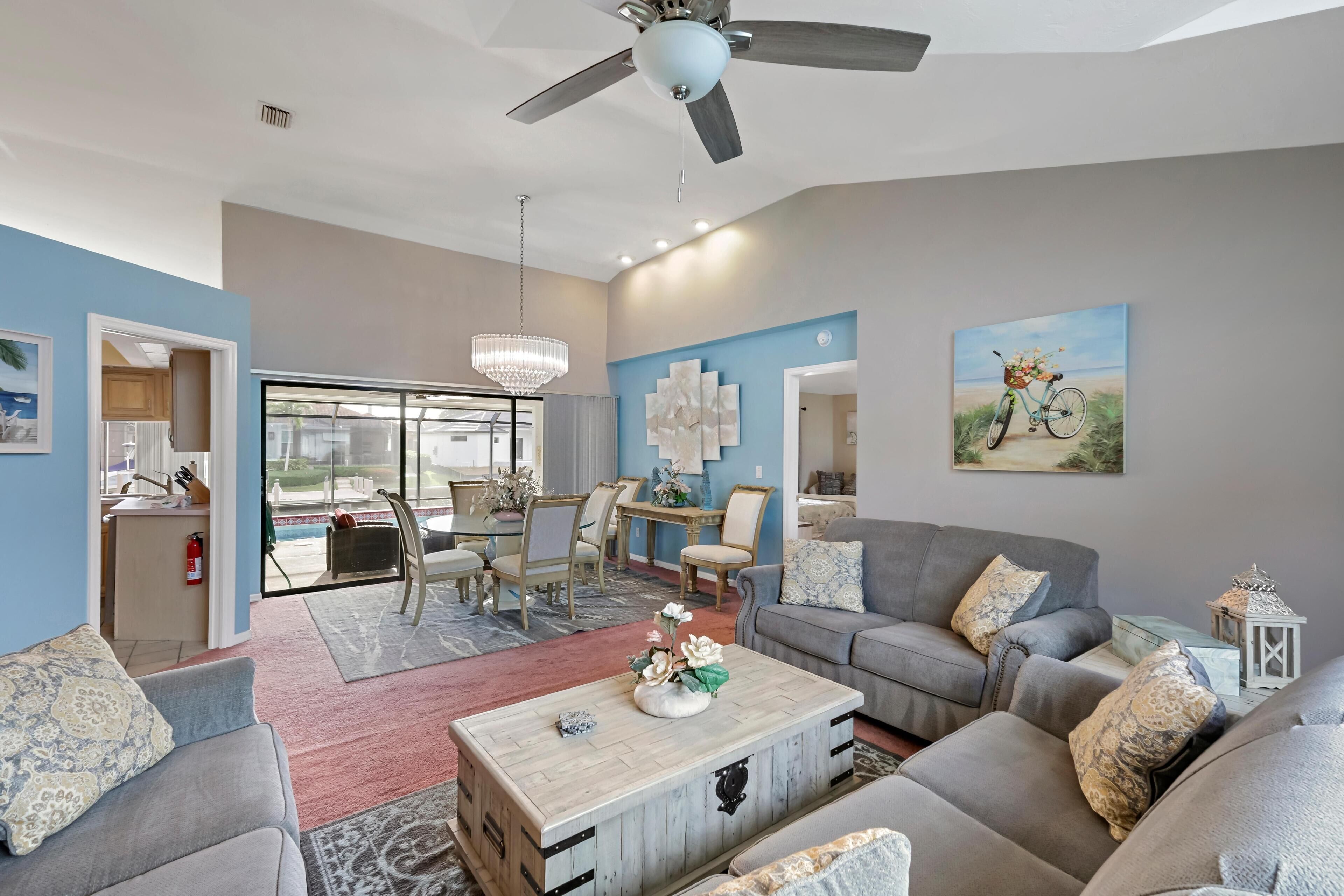 A spacious living room with a grey sofa, a wooden coffee table, and a chandelier. There is a dining area with a table and chairs, and a painting of a bicycle on the wall.