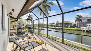 A sunlit patio with outdoor furniture, a view of a water body, and residential buildings in the background. - Quiet Waterfront Home With Pool, Dock and Bicycles (Marco Island)