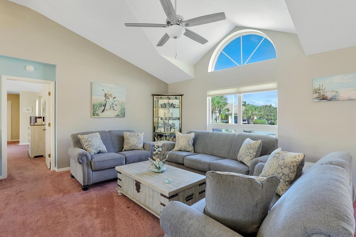 A spacious living room with a gray sectional sofa, a wooden coffee table, and a ceiling fan. There is a large window providing natural light and a view of greenery outside.