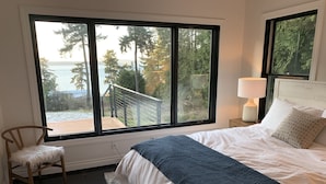 Un dormitorio con un ventanal grande, una cama con ropa de cama blanca, una silla de madera y una mesita de noche con una lámpara.