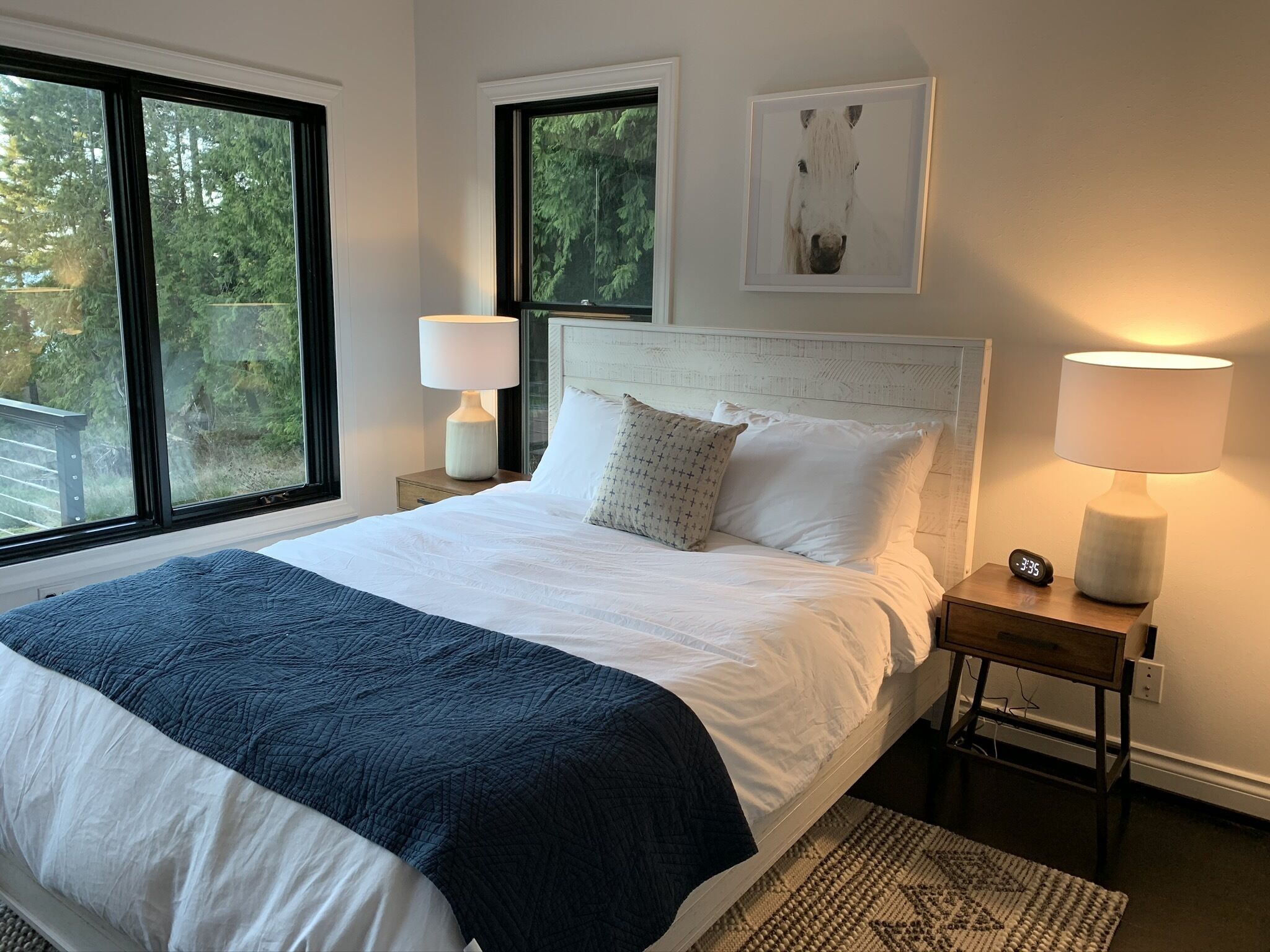 A neatly made bed with a blue blanket, two bedside lamps, a clock, and a framed picture on the wall.