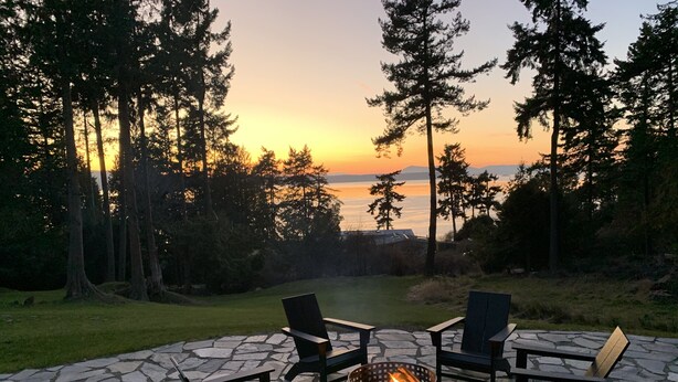 Un espacio al aire libre con zona de fogata rodeada de sillas Adirondack, frente a un paisaje de árboles y un atardecer sobre un cuerpo de agua.