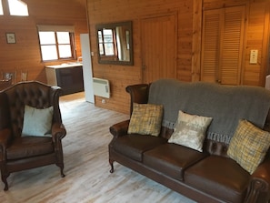 A wooden interior with a brown leather sofa and armchair, a herringbone-patterned throw, and a mirror on the wall.