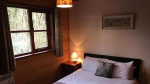 A wooden room with a bed, a nightstand with a lamp, a window with curtains, and a framed picture on the wall. - Shrew Lodge Hot Tub (Carmarthen)