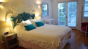 A bedroom with a large bed, a nightstand, a red rug, a white chair, and a window with blinds. - Beverly Hills Celebrity Home (Los Angeles)