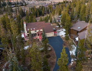 A large, two-story house with a red roof and a chimney, surrounded by dense forest and a small stream.