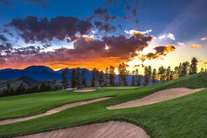En golfbana med välskött gräs, sandbunkrar och en vacker bakgrund av berg och träd under solnedgången.