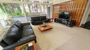 Uma sala de estar moderna com um sofá de couro, uma mesa de centro com uma planta, uma TV sobre um suporte de madeira e uma janela grande com vista para árvores.