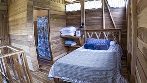 A wooden cabin room with a bed, a shelf with folded towels, and a window with a wooden ladder. - Oski Lodge  (Guayabo)
