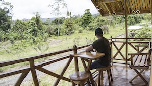 A wooden deck with a person seated at a table, overlooking a lush landscape with trees and a distant mountain. - Oski Lodge  (Guayabo)