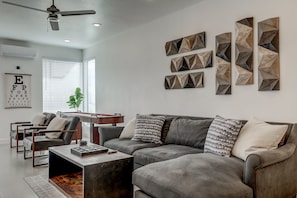 A modern living room with a grey sectional sofa, a coffee table, and a ceiling fan. There are abstract wall art pieces and a framed eye chart on the wall. - Pool Heated All Year | Blazing Fast Internet | #17 (Saint George)
