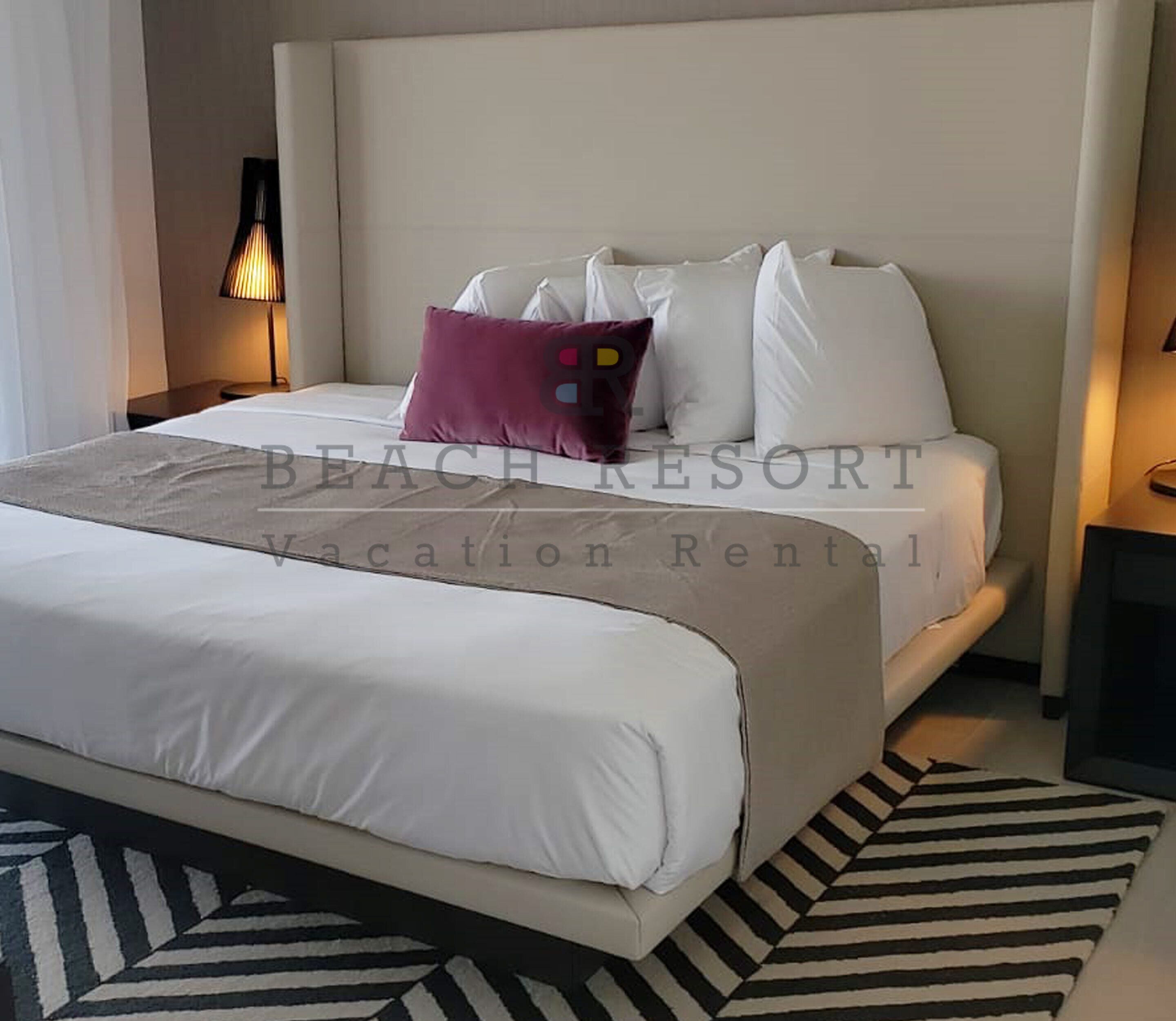 A neatly made bed with white linens and a single purple pillow, a striped rug, and a wall-mounted lamp.
