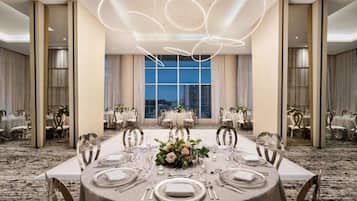 A formal dining room with round tables, white tablecloths, and a city view through large windows.