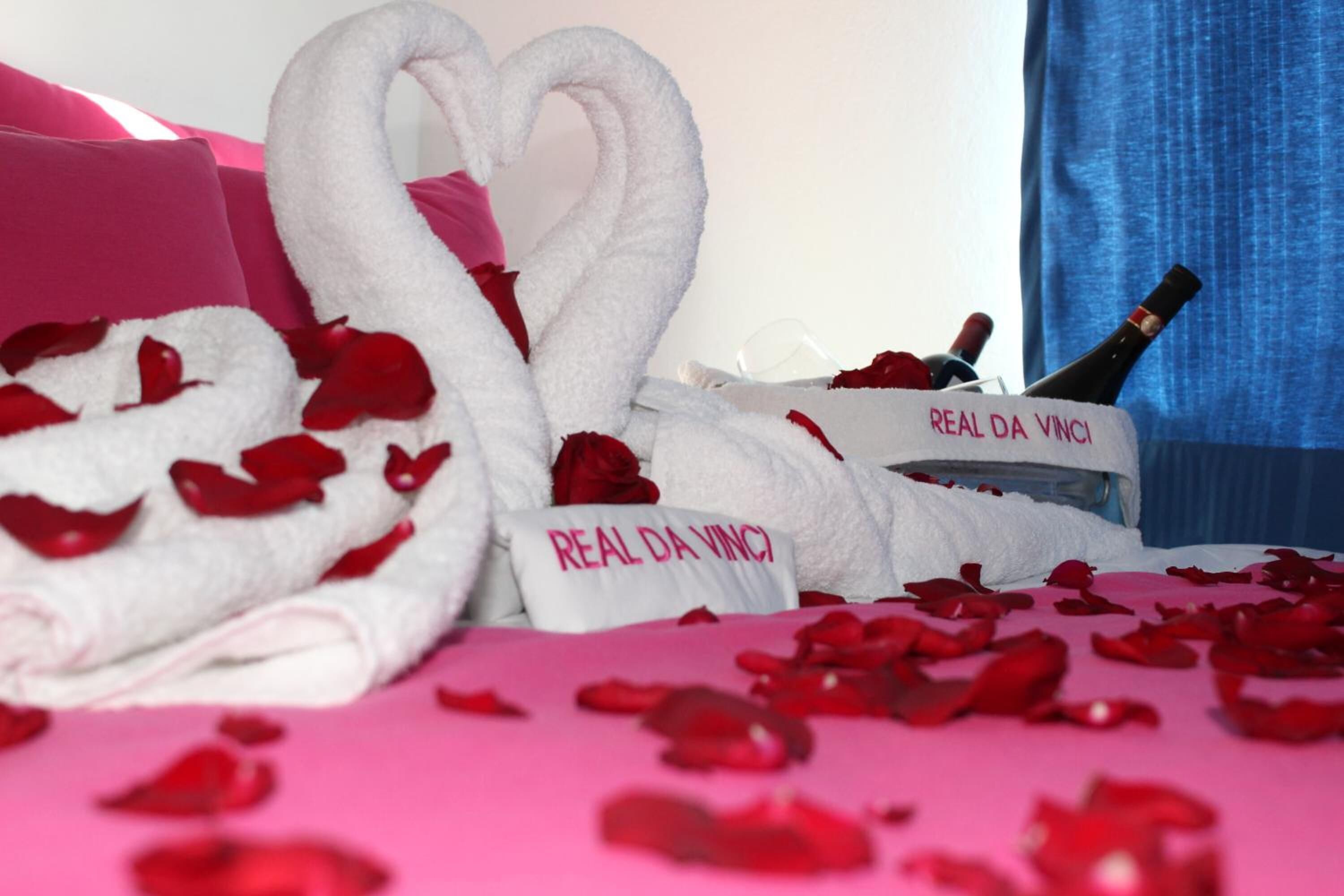 A bed with rose petals and two white towels shaped like swans with "REAL DA VINCI" printed on them.