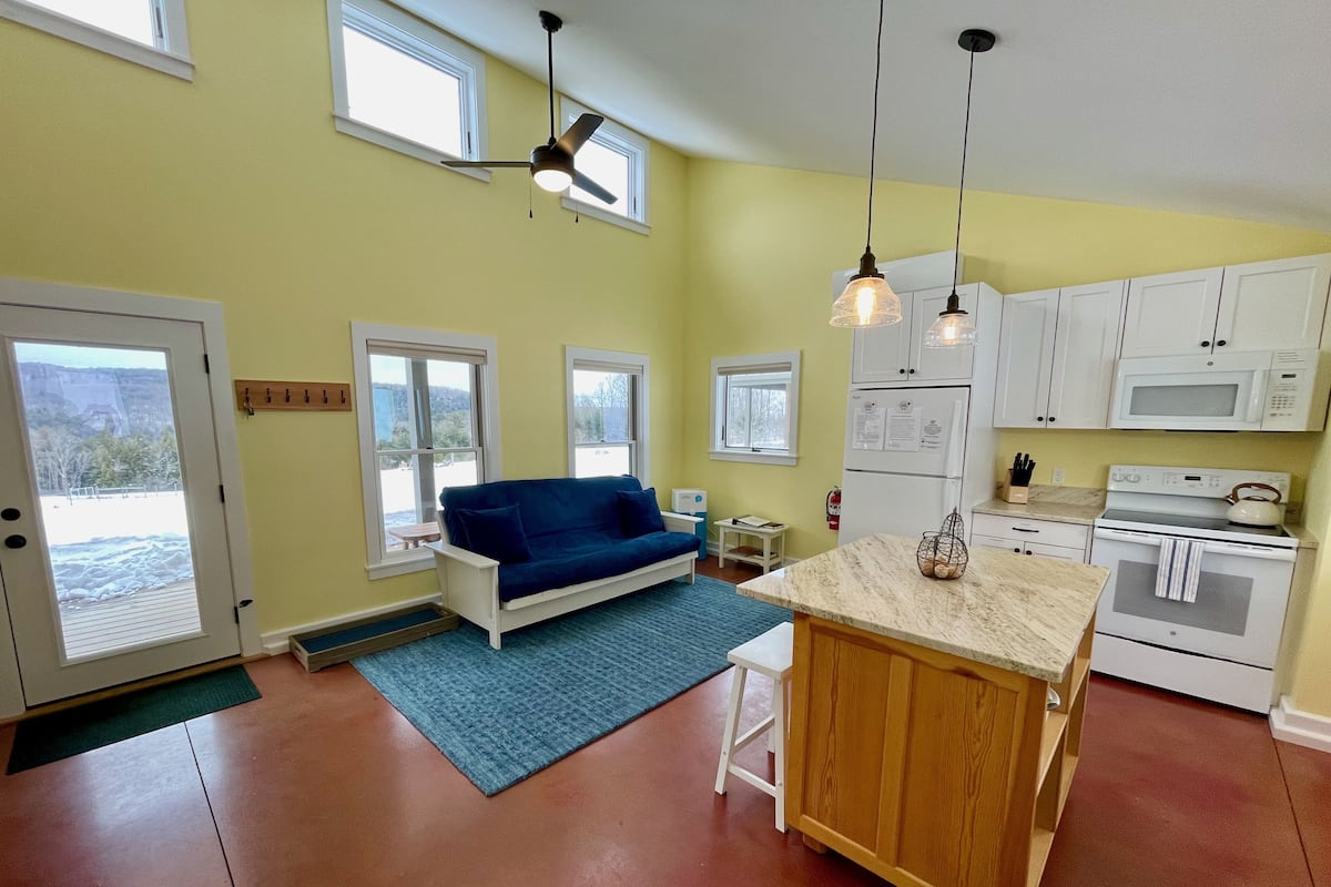 A compact kitchen with white cabinets, a built-in oven, and a microwave. There is a blue sofa and a small table in the living area.