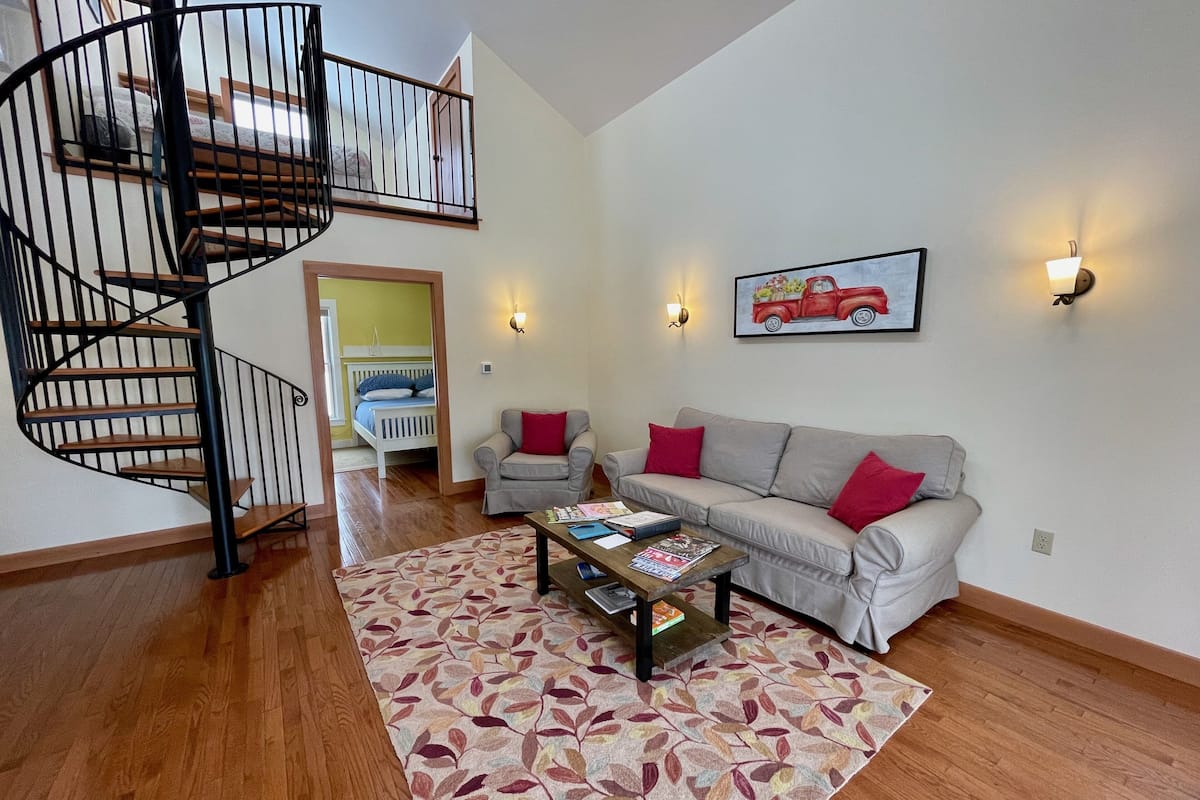 A spacious living room with a wooden floor, a grey sofa with red cushions, a small armchair, a coffee table with magazines, a spiral staircase, and a painting of a red car on the wall.
