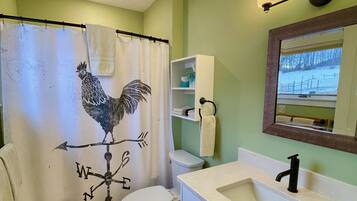 Casa de banho com lavatório branco, sanita, cortina de chuveiro com padrão de galo, espelho e prateleira com produtos de higiene pessoal.