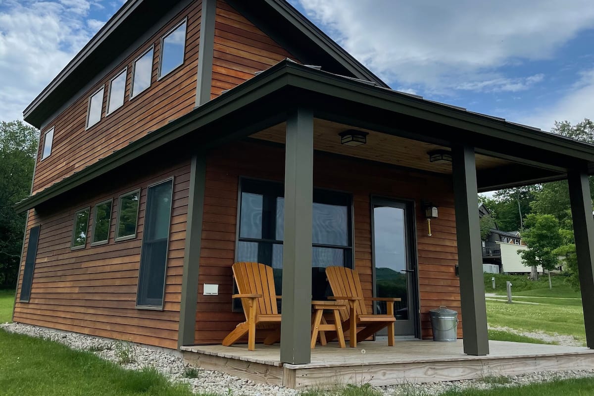 A two-story wooden house with a covered porch and two Adirondack chairs.