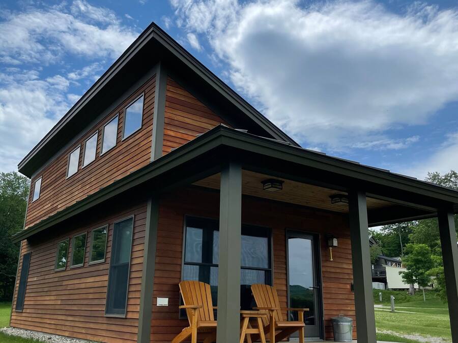 A two-story wooden house with a covered porch and two Adirondack chairs.