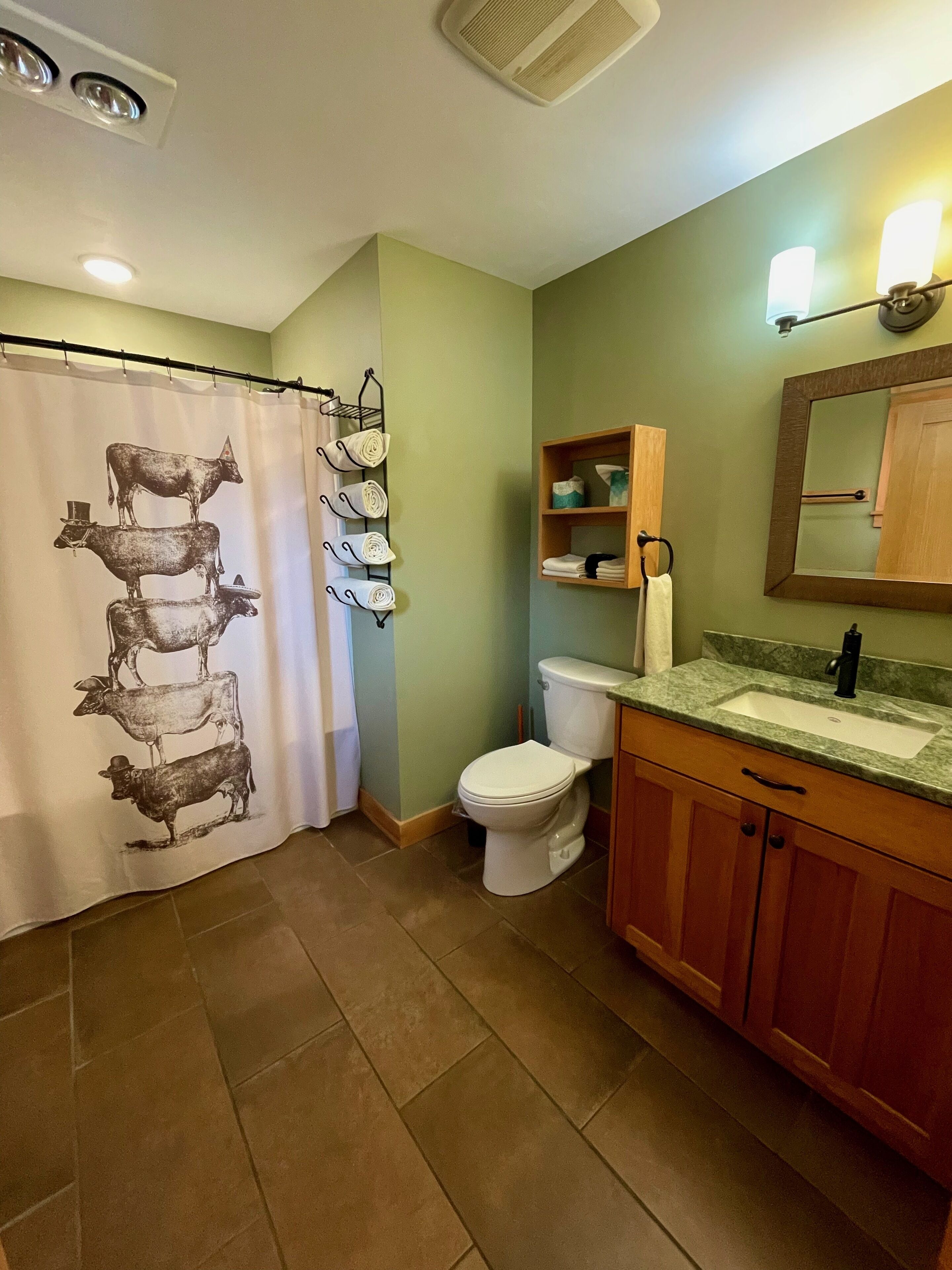 A bathroom with a shower curtain featuring cow illustrations, a toilet, a sink with a mirror above it, and wooden cabinets.