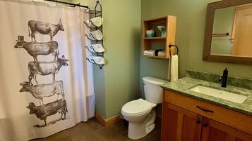 A bathroom with a shower curtain featuring cow illustrations, a toilet, a sink with a mirror above it, and wooden cabinets.