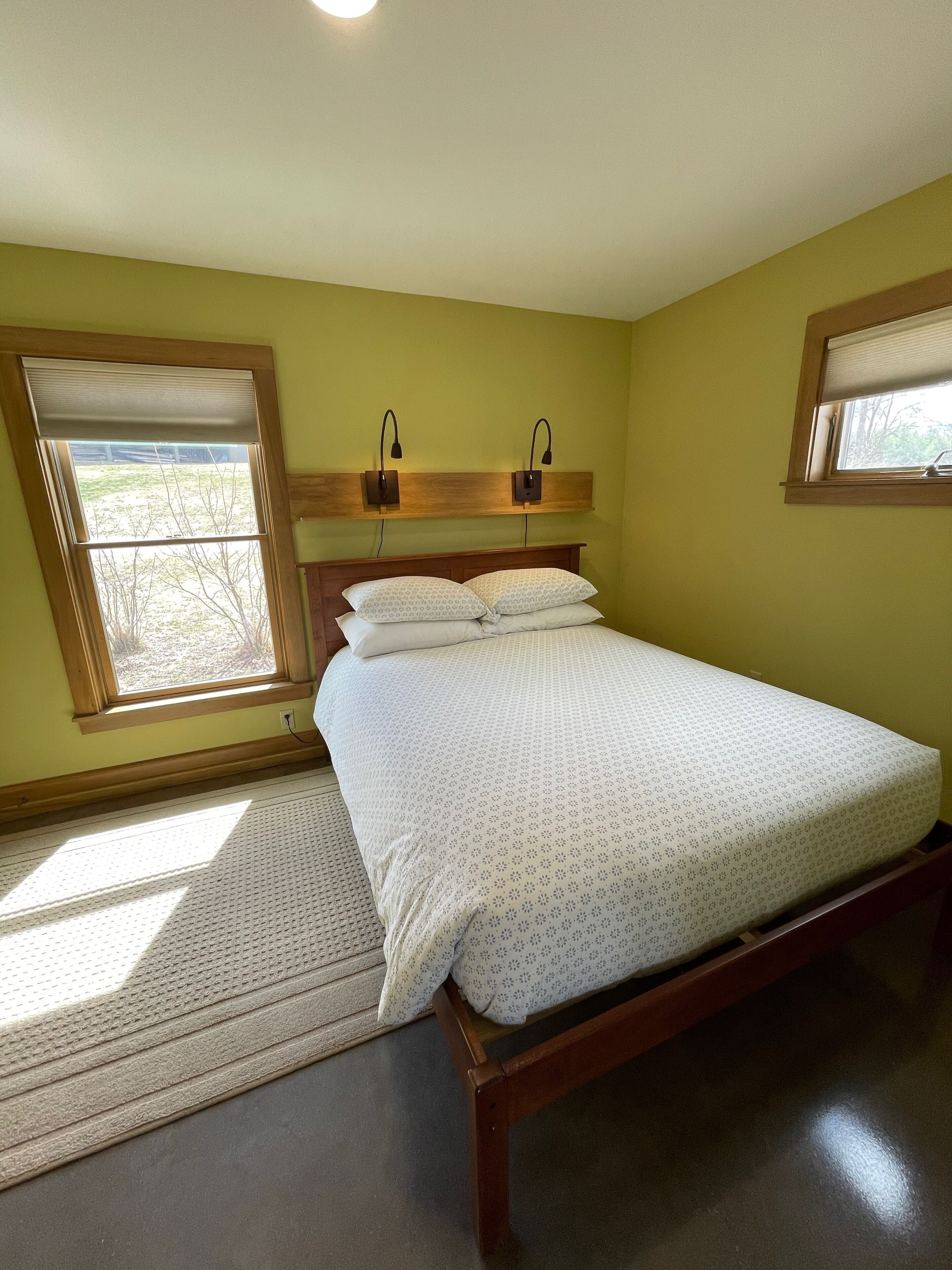 A bedroom with a bed, two bedside lamps, a window with blinds, and a view of the outdoors.