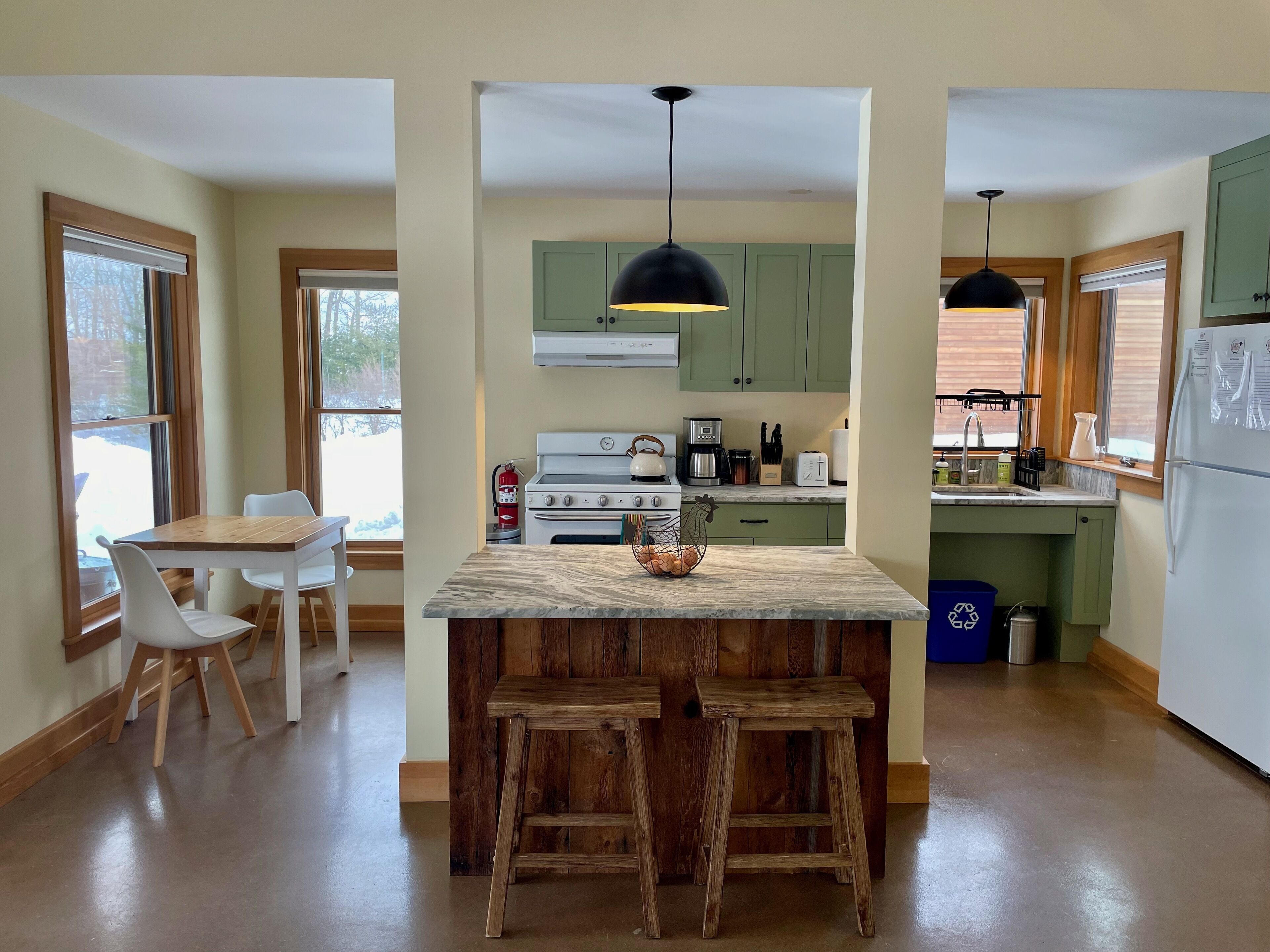 A kitchen with a central island, wooden stools, a stove, a sink, and green cabinets.
