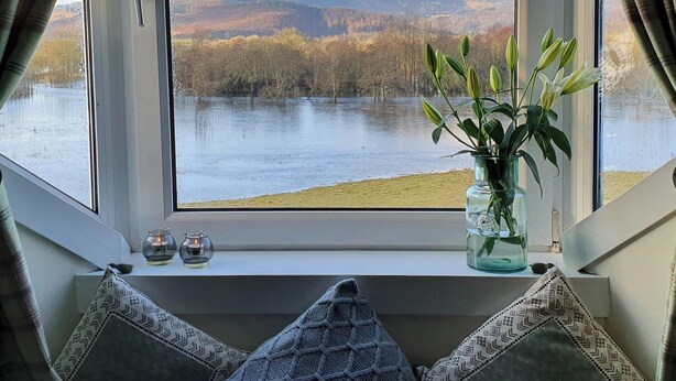 A window with a view of a lake and mountains, a vase with flowers, and two candles on the windowsill.