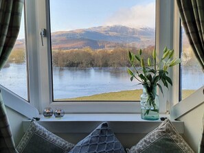 Una ventana con vista a un lago y montañas, un florero con flores y dos velas en el alféizar.