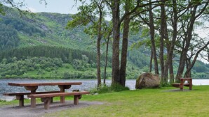 A picnic area with benches and a table by a lake, surrounded by trees and hills.