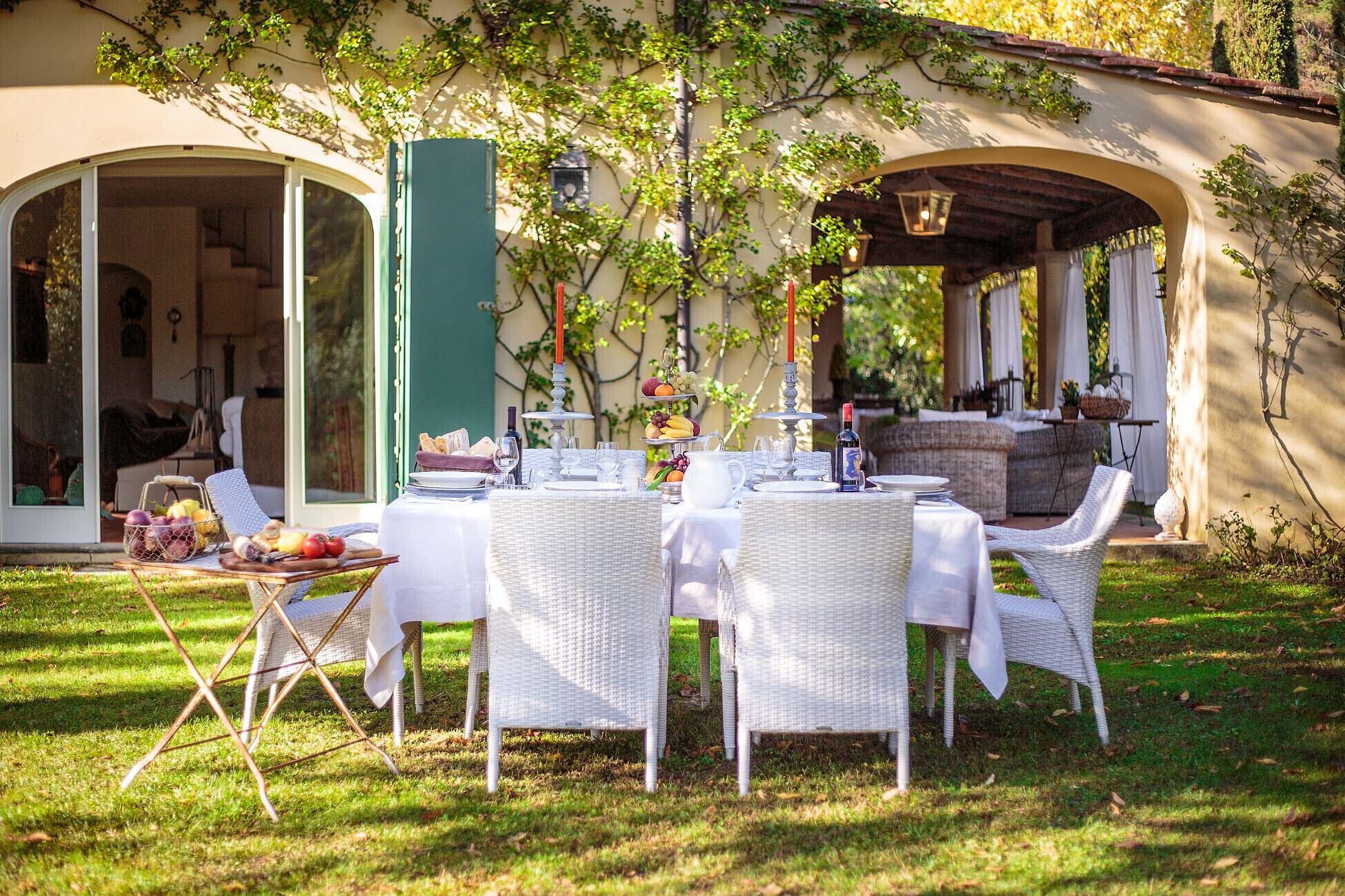 An outdoor dining area with wicker chairs and tables, a buffet with fruits and bread, and a covered seating area with a view of the garden.