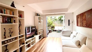 Un salon moderne avec un canapé blanc, une bibliothèque remplie de livres et d’objets décoratifs, un téléviseur à écran plat et un plancher en bois.