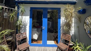 Une maison à deux étages avec une porte et des fenêtres bleues, une terrasse en bois et diverses plantes en pot.
