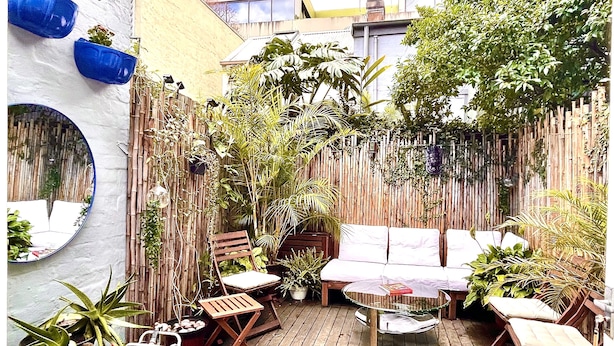 Une terrasse en bois avec une table ronde en verre et des chaises, entourée de plantes en pot et d’une clôture en bambou.