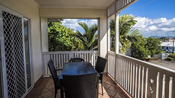 A balcony with a table and chairs, surrounded by a white railing, overlooking a view of palm trees and buildings. - Cairns City Garden Apartment (Parramatta Park)