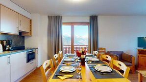 Un comedor con una mesa dispuesta para cuatro, una cocina con electrodomésticos modernos y vistas a montañas a través de amplios ventanales.