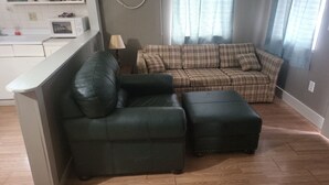 Una sala de estar con un sofá verde oscuro, un otoman a juego y un sofá a cuadros. Una lámpara está sobre una mesita auxiliar y hay un microondas en la encimera.