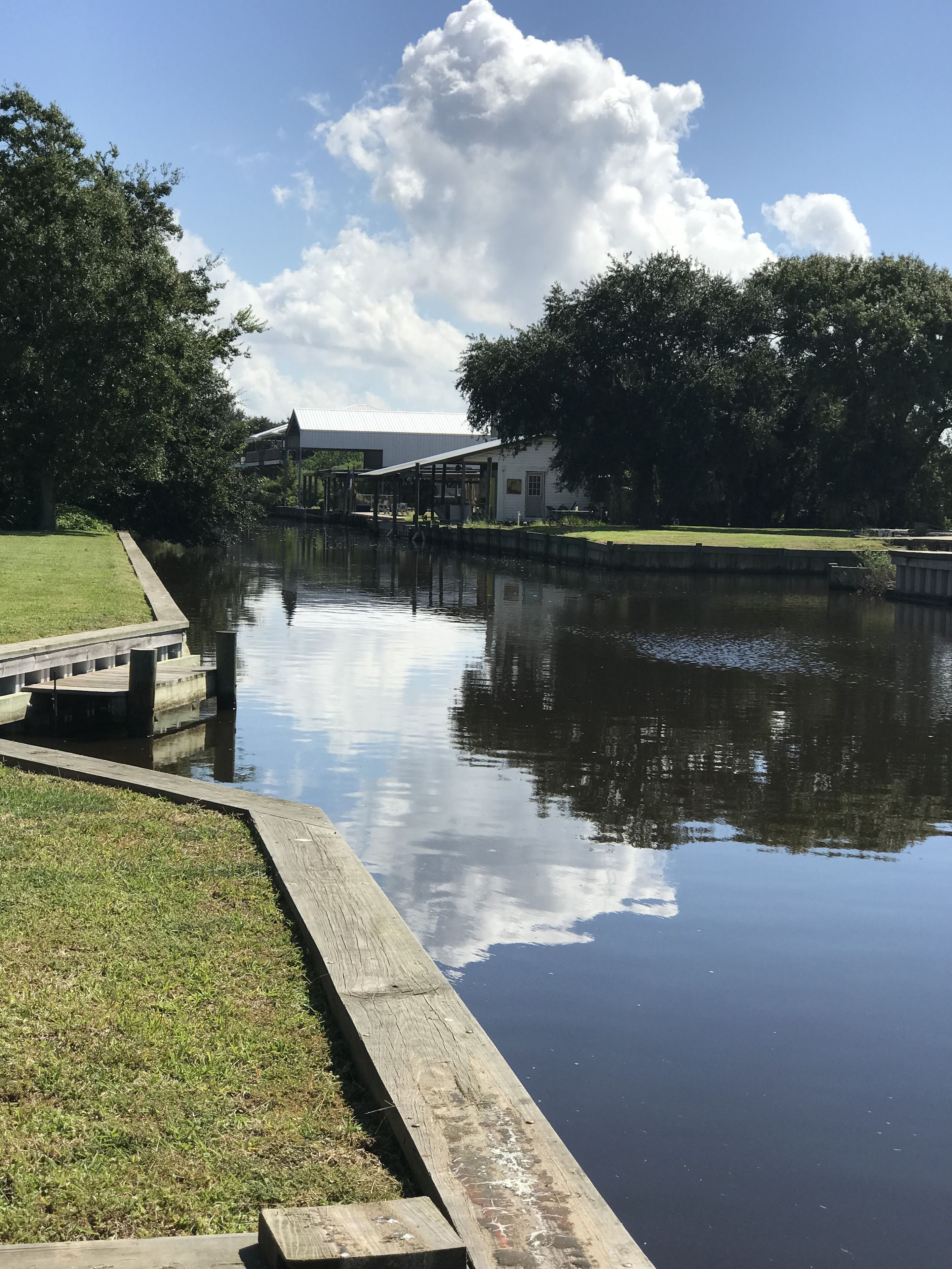 A calm water body with a wooden dock, a grassy area, and a building with a white roof in the background.