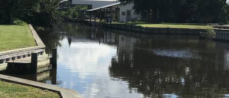 A calm water body with a wooden dock, a grassy area, and a building with a white roof in the background.