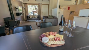 A dining area with a wooden board of cheese, grapes, nuts, and crackers. There are two wine glasses and a bottle of wine on the table.