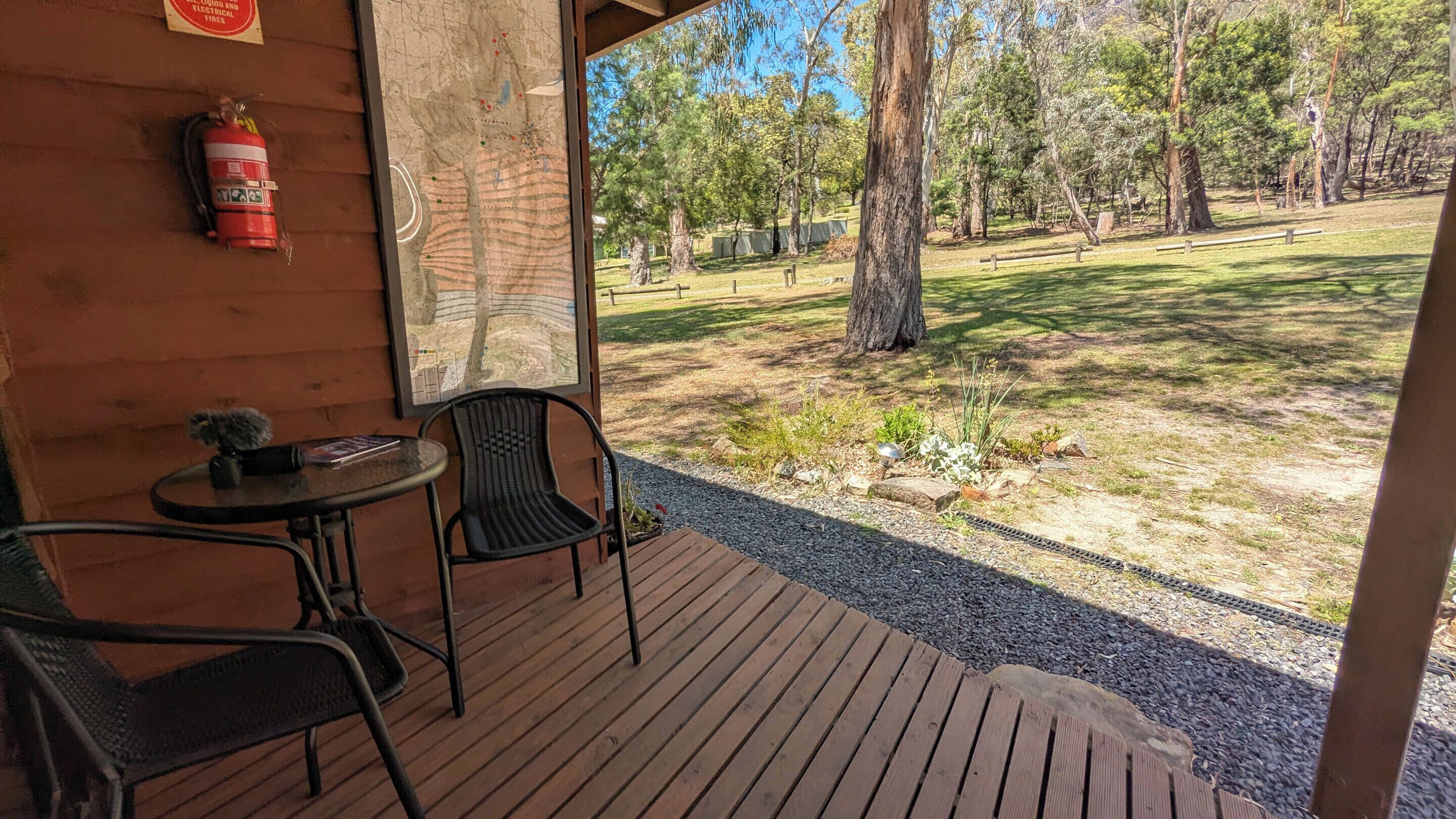 Una terraza de madera con una mesa y sillas, un extintor y vistas a una zona boscosa.