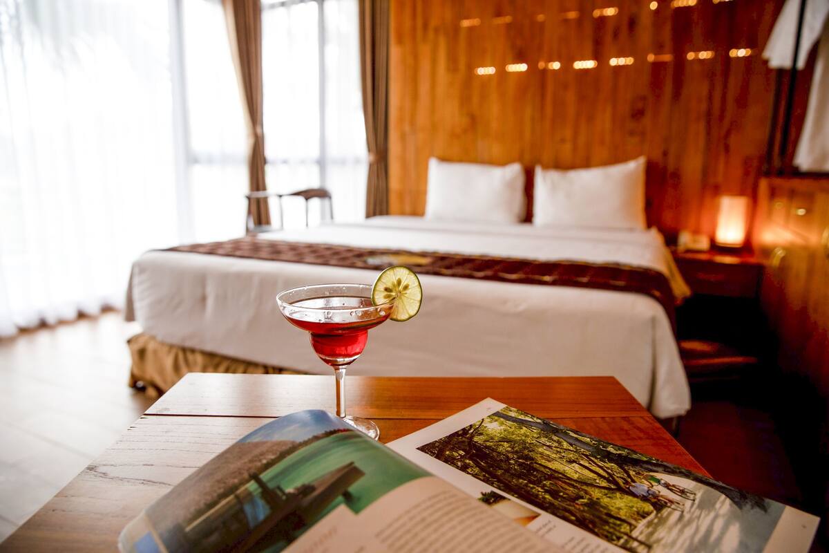 A hotel room with a bed, a table with a drink and a magazine, and a window with curtains.