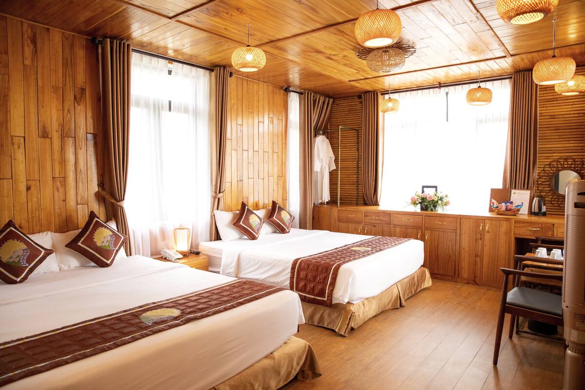 A hotel room with two beds, wooden walls, a wooden ceiling, and a wooden floor. There is a desk with a chair and a window with curtains.