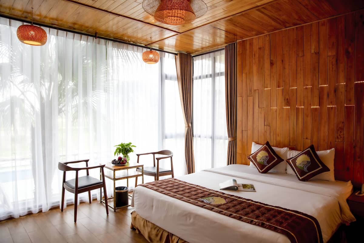 A room with a large bed, wooden walls, a desk with a chair, and a window with sheer curtains.
