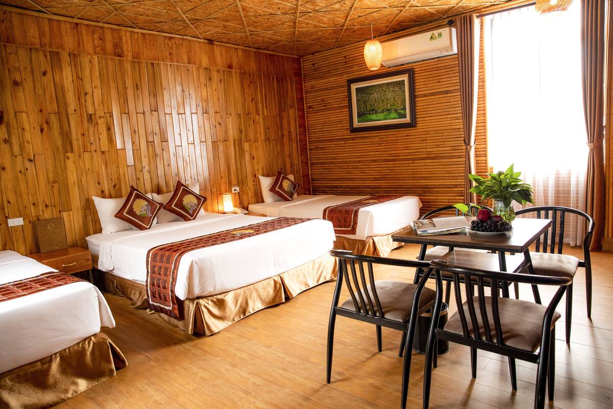 A room with wooden walls, two beds with white bedding, a dining table with chairs, and a potted plant.