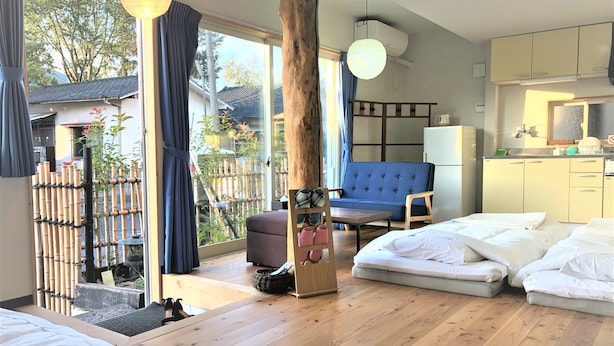 A modern room with a bed, a sofa, a dining table, and a view of a bamboo fence and trees outside.