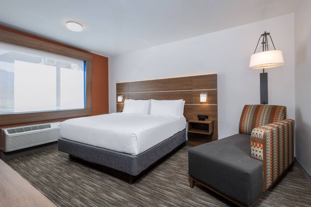 A hotel room with a large bed, a bedside table, a chair, a lamp, and a window with blinds.